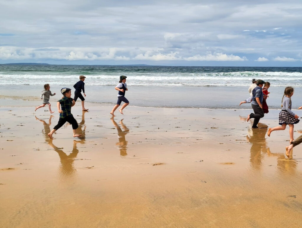 A group of children in historical costumes running along a beach shore.