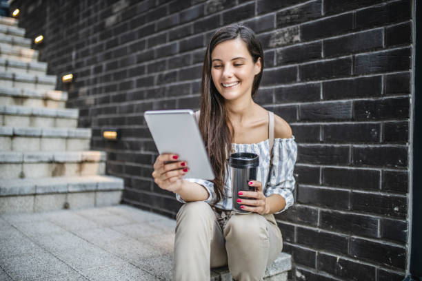 Young woman sitting on stairs, using digital tablet, drinking coffee, smiling and taking a break