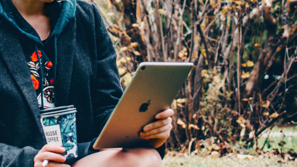 Photo of young woman in a park reading on an iPad and holding a coffee.