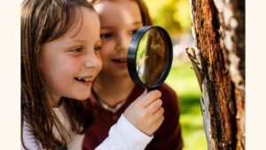 Two girls look at insects on a tree with a magnifying glass.