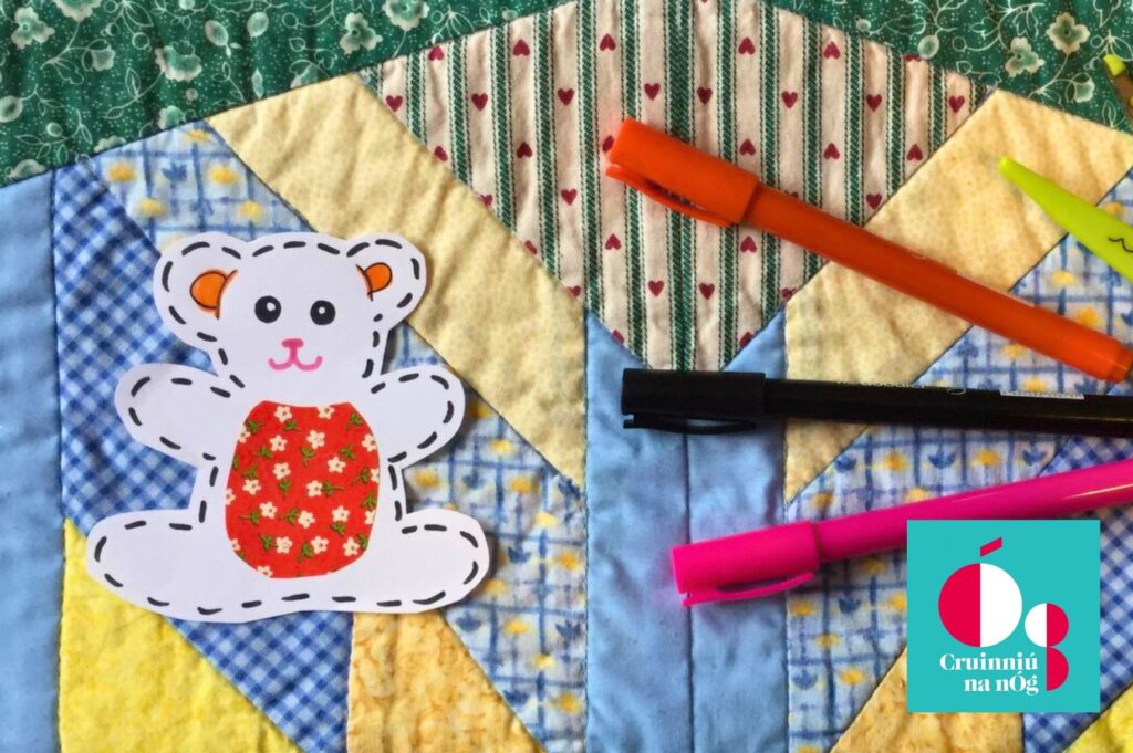 A patchwork quilt with a white teddy surrounded by patchwork of various sizes and shapes. There are three markers resting on the quilt in colours of red, pink and black.