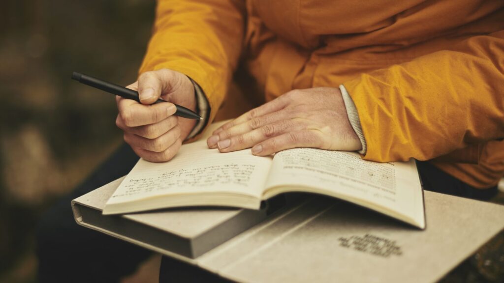 A person in a yellow jacket sits with a pen in their hand. There is an open writing book on the table with words written on a page.