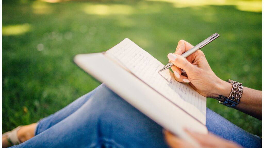 A photograph of a person sitting outside on the grass with an open journal on their lap. They are writing in the journal with a pen.