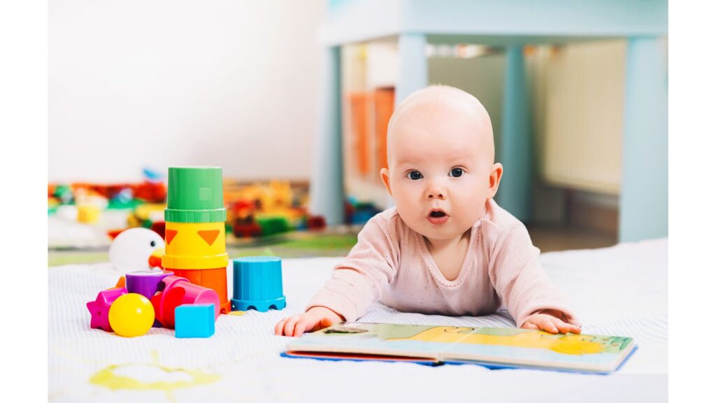 A baby lying on his/her tummy on the floor with a book in front. Some colourful toys are beside the baby.